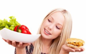 thoughtful young woman holding a hamburger with chicken and plate with vegetables, isolated against white background